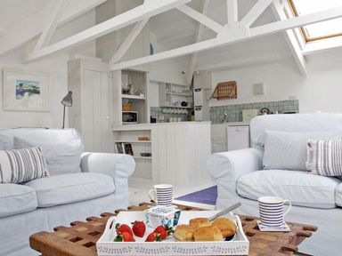 A living area with white sofas and striped pillows a wooden coffee table with food and drinks and a kitchen in the background at The Net Loft in Mousehole