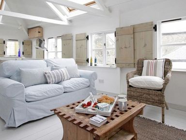 A sitting area with a sofa and wicker chair around a wooden coffee table in a room with windows and wooden shutters at The Net Loft in Mousehole
