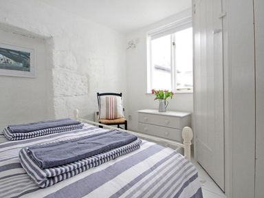 A bedroom with a bed with striped bedding two folded towels a chair with a cushion a dresser with flowers and a window at The Net Loft in Mousehole