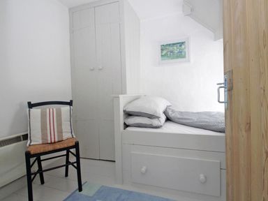 A small bedroom with a single bed with striped bedding a wooden chair with a striped cushion a closed wardrobe and a framed picture on the wall at The Net Loft in Mousehole