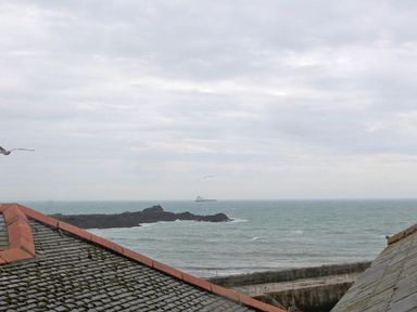 A view of the sea with a rocky outcrop and a ship on the horizon seen between two sloping tiled roofs