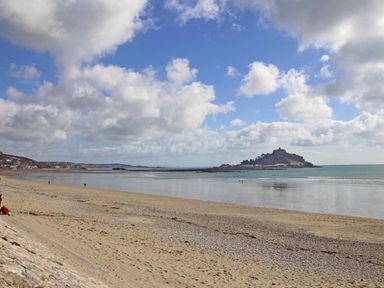 A sandy beach with a rocky shoreline and an island with a castle in the distance under a partly cloudy sky at The Net Loft in Mousehole