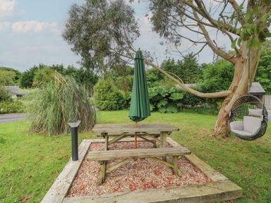 An outdoor area with a wooden picnic table and bench under a closed green umbrella near a tree with a hanging chair at The Stables in Ruthvoes near St Columb Major