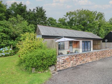 A small house with a patio umbrella and stone wall surrounded by trees and bushes at The Stables in Ruthvoes near St Columb Major