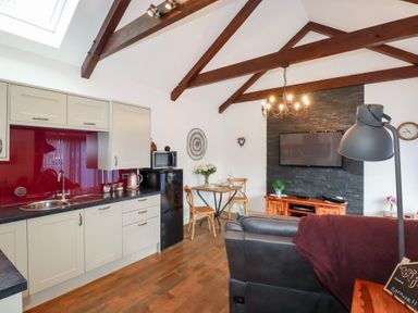 An open plan kitchen and living area with wooden beams a dining table a leather sofa and a wall mounted television at The Stables in Ruthvoes near St Columb Major