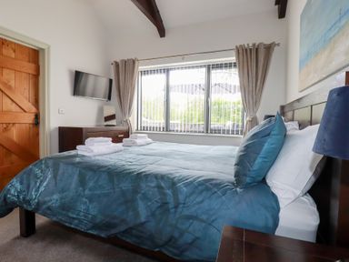 A bedroom with a blue bedspread and stacked towels on the bed a wooden door a wall-mounted television and a window with curtains at The Stables in Ruthvoes near St Columb Major