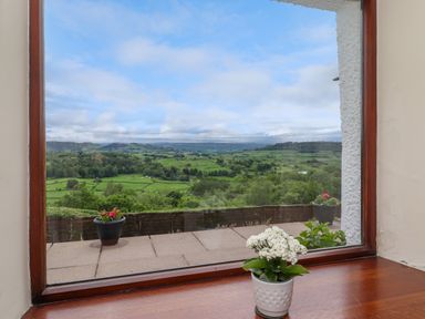 A window with a potted white flower on the sill overlooking a patio with two potted plants and green fields at Addyfield Farmhouse in Cartmel Fell near Bowland Bridge