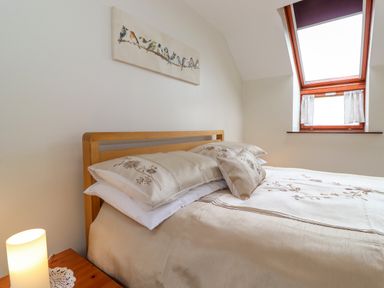 A bedroom with a bed with beige floral bedding a wooden bedside table with a lamp and a window with white curtains at Addyfield Farmhouse in Cartmel Fell near Bowland Bridge