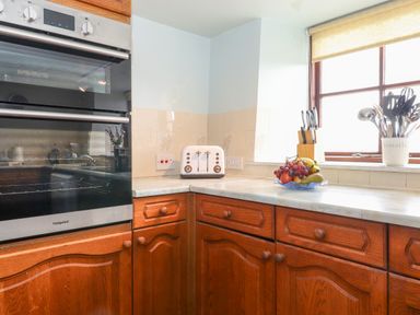 A kitchen corner with wooden cabinets a double oven a white toaster a fruit bowl and a utensil holder near a window at Addyfield Farmhouse Cartmel Fell near Bowland Bridge