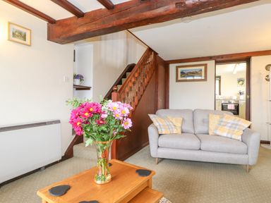 A living room with a gray sofa and a wooden coffee table with flowers near a wooden staircase at Addylea Cottage in Cartmel Fell near Windermere