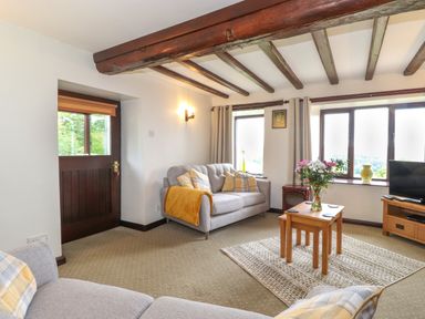 A living room with wooden beams on the ceiling two sofas a coffee table with flowers and a television on a wooden stand at Addylea Cottage in Cartmel Fell near Windermere