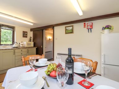 A dining table set with bowls glasses fruit and a wine bottle in a kitchen with cabinets a microwave and a refrigerator at Addylea Cottage in Cartmel Fell near Windermere