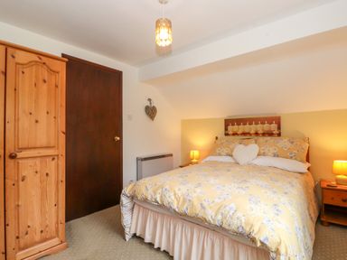 A bedroom with a double bed having floral bedding two nightstands with lamps wooden wardrobe and a closed dark door at Addylea Cottage in Cartmel Fell near Windermere