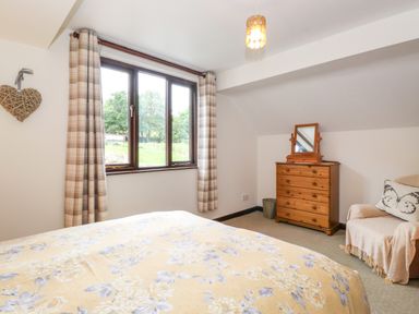 A bedroom with a bed covered in floral bedding a wooden dresser with a mirror a window with plaid curtains and a chair with a butterfly cushion at Addylea Cottage in Cartmel Fell near Windermere