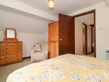 A bedroom with a chest of drawers with a mirror an armchair with a butterfly cushion a wooden wardrobe and a bed with a floral bedspread at Addylea Cottage in Cartmel Fell near Windermere
