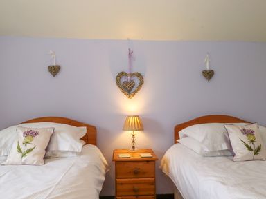 A bedroom with two single beds with white bedding and floral cushions a wooden nightstand with a lamp and three woven heart decorations on the wall at Addylea Cottage in Cartmel Fell near Windermere