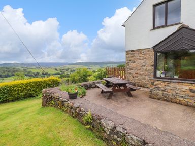 An outdoor patio with a wooden picnic table next to a stone and white house overlooking a green landscape at Addylea Cottage Cartmel Fell near Windermere