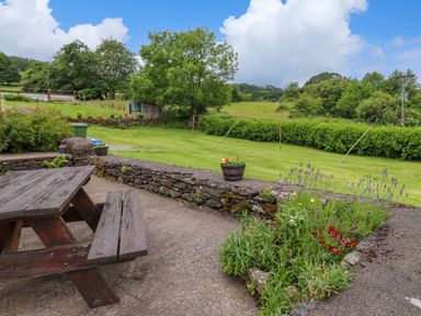 An outdoor patio with a wooden picnic table and bench next to a stone wall with flowerbeds and green lawn beyond at Addylea Cottage in Cartmel Fell near Windermere