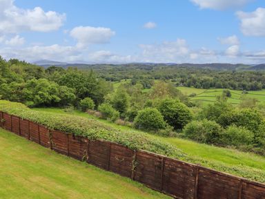 A wooden fence with grass and trees on a hillside overlooking fields and distant hills at Addylea Cottage in Cartmel Fell near Windermere