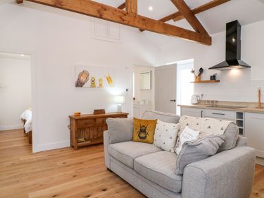 A living room with a gray sofa and kitchen area with wooden beams on the ceiling at Pear Tree Cottage in St Mellion near St Dominick