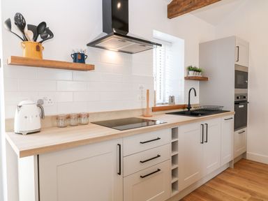 A kitchen with white cabinets wooden countertops and shelves a black stovetop a white kettle and utensils in jars at Pear Tree Cottage in St Mellion near St Dominick