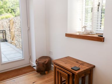 A corner with nested wooden tables a brown animal-shaped stool a window with blinds and a dog figurine on the sill and a glass door leading outside at Pear Tree Cottage in St Mellion near St Dominick