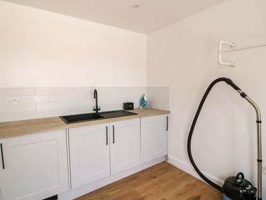 A utility room with white cabinets a black sink and faucet an iron and a vacuum cleaner on a wooden floor at Pear Tree Cottage in St Mellion near St Dominick