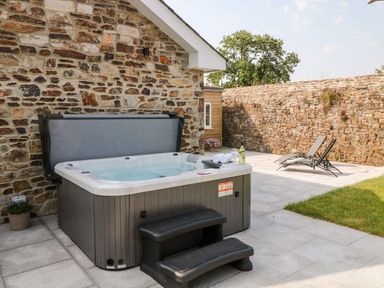 An outdoor patio area with a hot tub and two lounge chairs next to a stone wall at Pear Tree Cottage in St Mellion near St Dominick