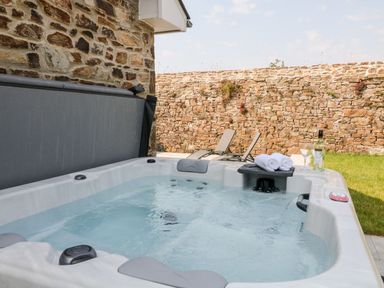 An outdoor hot tub with water jets next to a stone wall with two lounge chairs and a bottle of wine with glasses at Pear Tree Cottage in St Mellion near St Dominick