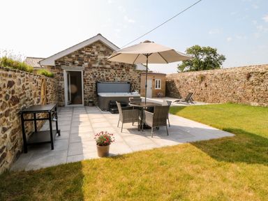 A patio with a table umbrella and chairs a hot tub a small black barbecue and garden chairs in a grassy yard at Pear Tree Cottage in St Mellion near St Dominick