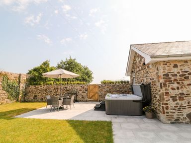 A patio with outdoor furniture and umbrella next to a hot tub by a stone building at Pear Tree Cottage in St Mellion near St Dominick