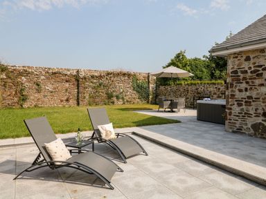 An outdoor patio with two lounge chairs and a table with a bottle and glasses next to a lawn and a stone wall at Pear Tree Cottage in St Mellion near St Dominick