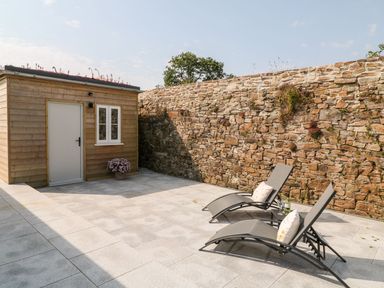 An outdoor patio area with two lounge chairs with cushions a small wooden shed a stone wall and a potted plant at Pear Tree Cottage in St Mellion near St Dominick