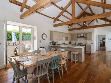 A kitchen with wooden beams and a dining table with chairs near open doors to an outdoor patio at Old Orchard Barn St Mellion near St Dominick