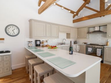 A kitchen with wooden stools at a white counter with a bottle and glasses and wooden beams on the ceiling at Old Orchard Barn St Mellion near St Dominick