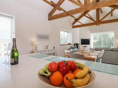 A dining area with a table set for six near a living room and a fruit bowl on a kitchen island at Old Orchard Barn in St Mellion near St Dominick