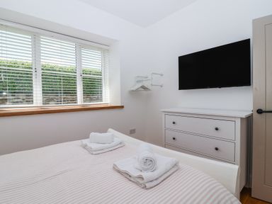 A bedroom with a bed with folded towels on it a window with blinds a white chest of drawers and a wall mounted television at Old Orchard Barn in St Mellion near St Dominick