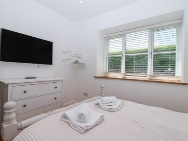 A bedroom with a bed, folded towels, a chest of drawers, wall-mounted television, window with blinds at Old Orchard Barn St Mellion near St Dominick