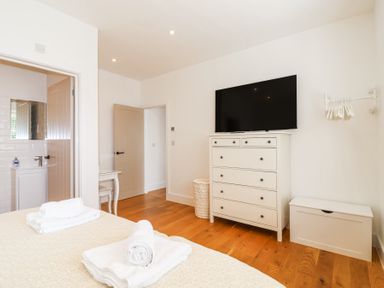 A bedroom with a bed and towels a white dresser with a TV on top a door open to a bathroom and wooden floor at Old Orchard Barn St Mellion near St Dominick