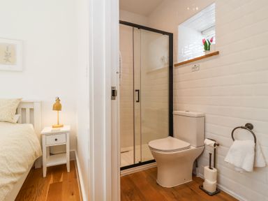 A bedroom with a bed and a nightstand with a lamp next to an adjoining bathroom with a toilet and a shower at Old Orchard Barn in St Mellion near St Dominick