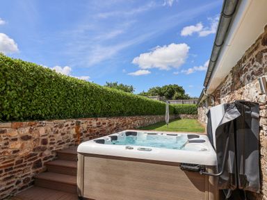 An outdoor hot tub next to a stone wall and green hedge under a blue sky at Old Orchard Barn St Mellion near St Dominick