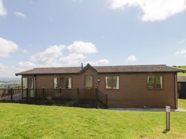 A brown wooden lodge with a railing on a grassy hill under a partly cloudy sky at Lodge 16 Whitsand Bay near Millbrook