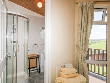 A bathroom with a corner shower and a small stool next to a window with curtains and a view of green fields at Lodge 16 Whitsand Bay near Millbrook