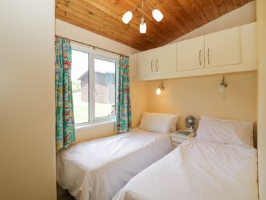 A bedroom with two single beds a window with colorful curtains and white cabinets above the beds at Lodge 16 Whitsand Bay near Millbrook
