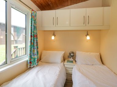A bedroom with two single beds a nightstand with a fan and book overhead cabinets and a window with a colorful curtain at Lodge 16 Whitsand Bay near Millbrook