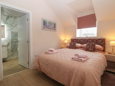 A bedroom with a double bed covered in pink bedding two folded towels on the bed side tables with lamps a window with a pink blind and an open door showing a bathroom with marble walls at Y Felin in Caernarfon