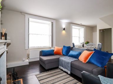 A living room with a gray sectional sofa with blue and orange cushions next to two windows with white shutters and a small dining table in the background at Carew House Cottage in St Day