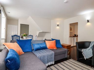 A living room with a grey sectional sofa with blue and orange cushions and a dining table with blue chairs at Carew House Cottage in St Day