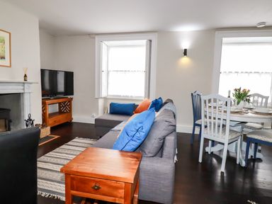 A living room with a gray sofa with blue and orange pillows a wooden side table a dining table with chairs and flowers on the table at Carew House Cottage in St Day
