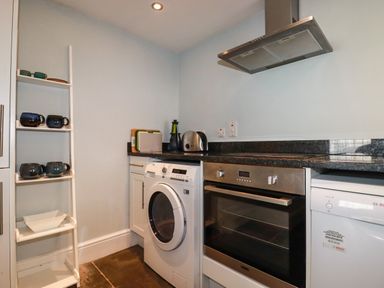 A kitchen area with a washing machine oven dishwasher countertop and shelves with mugs at Carew House Cottage in St Day